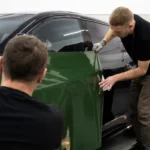 Professional automotive detailer carefully applying paint protection film to the bonnet of a luxury car in a clean detailing studio.